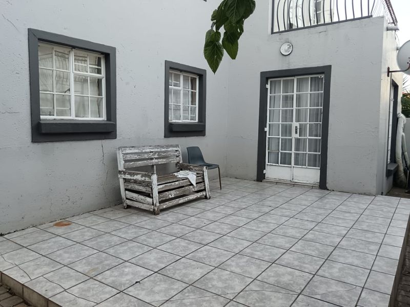 A paved outdoor patio area featuring tiled flooring, french doors, and casement windows, showing signs of wear on the exterior wall plaster.