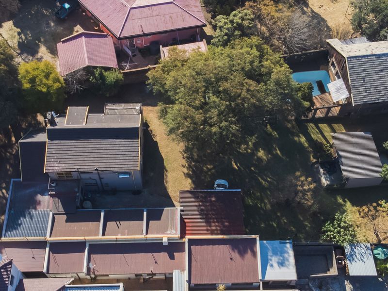 Aerial view of the property showing multiple structures, a swimming pool, mature trees, and driveway access within a residential setting.
