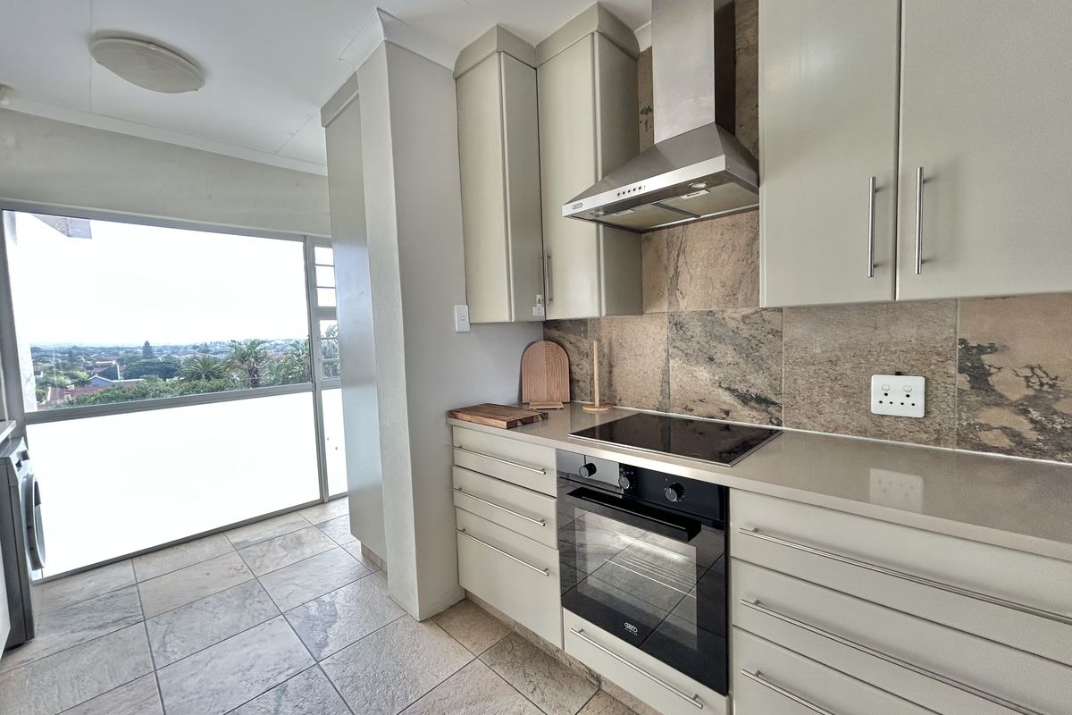 Modern kitchen featuring neutral cabinetry, stone countertops, built-in oven and hob, tiled splashback, and direct access to a balcony with scenic views.