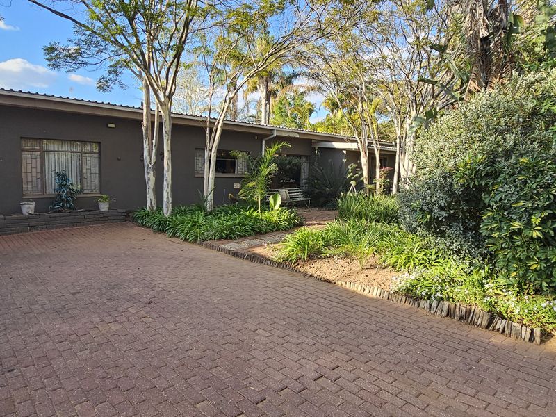 A view of the front exterior of the house featuring a paved driveway, established garden beds, and mature trees.