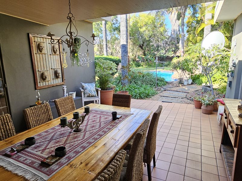 A covered patio area featuring tiled flooring, overlooking a lush garden and swimming pool, ideal for outdoor dining and entertaining.