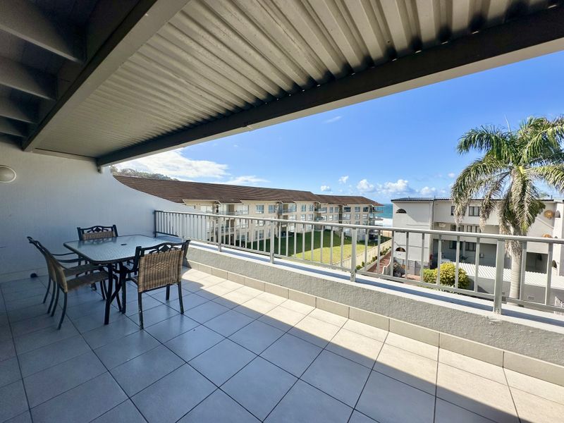 A spacious covered balcony featuring tiled flooring, a metal balustrade, and views of the surrounding complex and distant ocean.