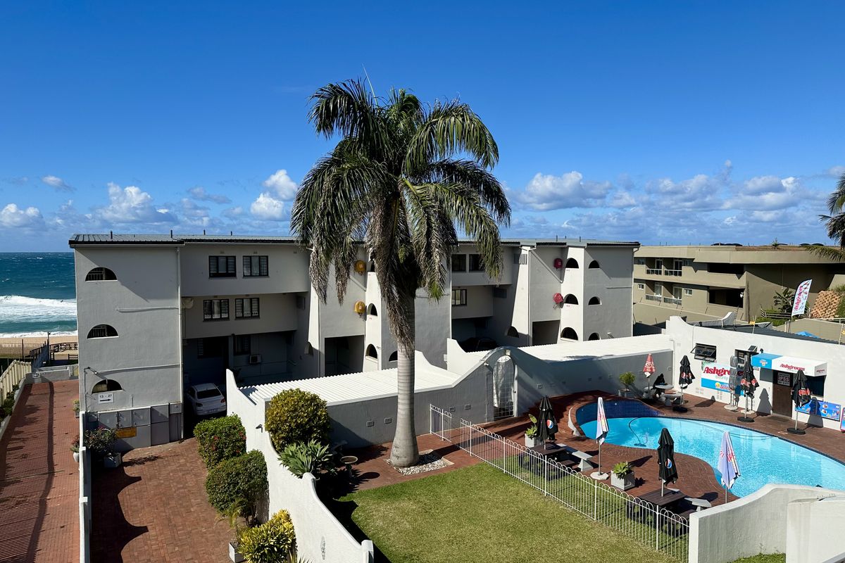 Elevated view of the apartment complex rear exterior, featuring a communal swimming pool, landscaped lawn, paved driveway, and direct proximity to the beach.