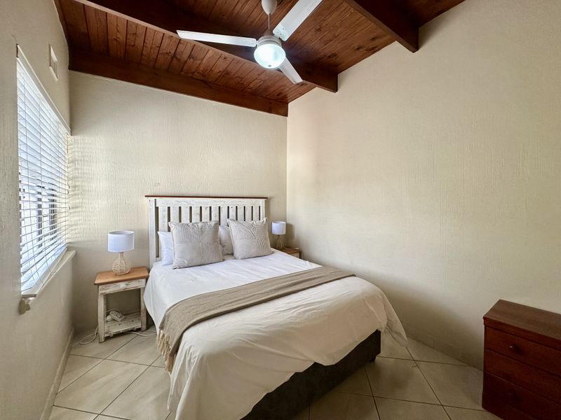 A cozy bedroom featuring a vaulted timber ceiling, ceiling fan, tiled flooring, and venetian blinds, offering a warm and functional living space.
