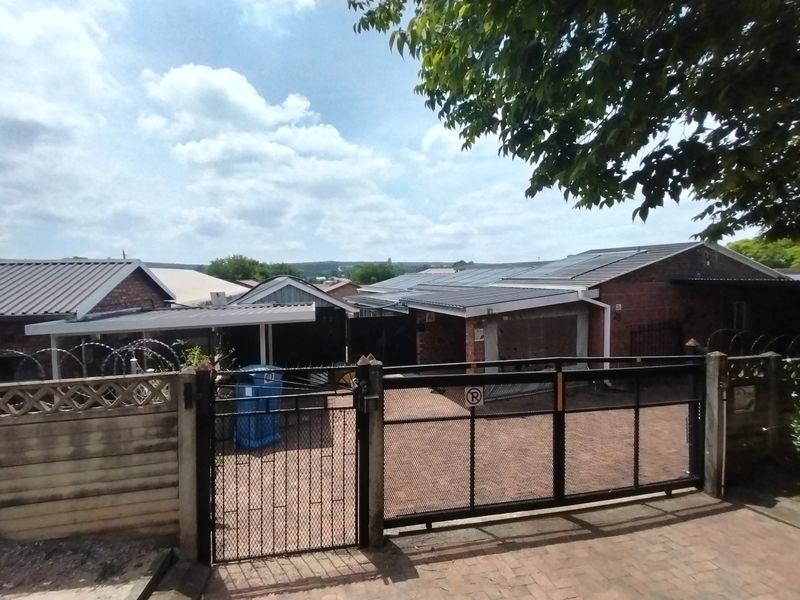Front view of a residential property featuring a brick facade, metal roofing with solar panels, a paved driveway, and a secure metal gate.