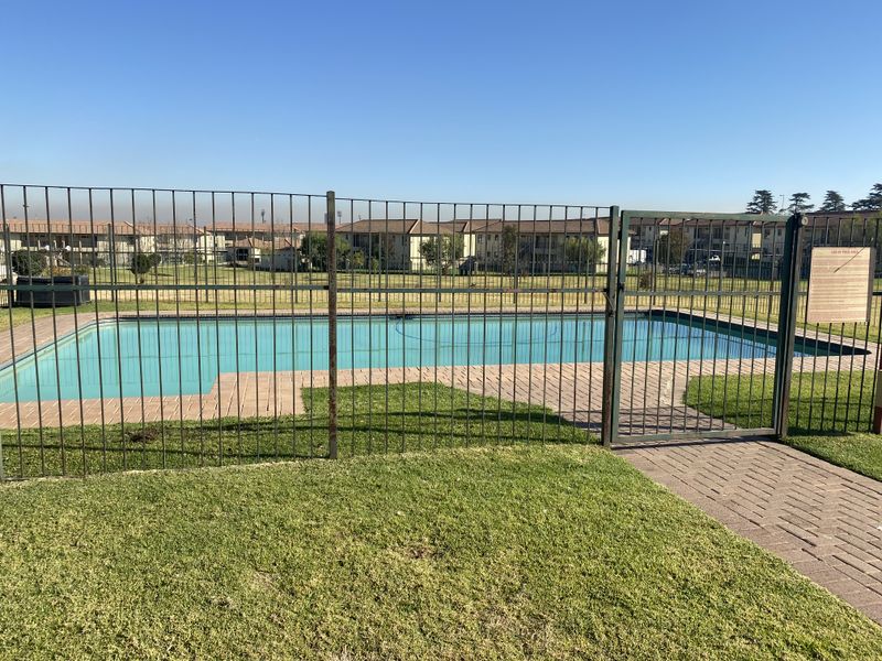 Communal swimming pool area featuring a paved surround, manicured lawn, and secure metal perimeter fencing within the townhouse complex.