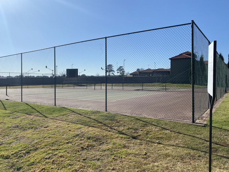 A well-maintained outdoor tennis court facility enclosed by high chain-link fencing, featuring a hard surface and integrated floodlighting.