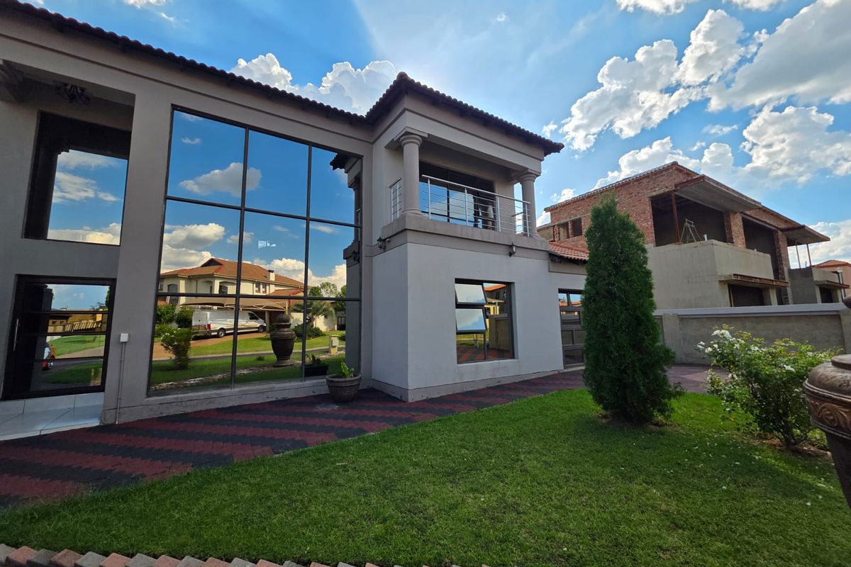 A modern double-story residential facade featuring extensive glass curtain walling, a covered balcony with balustrades, and a well-maintained garden with paved walkways.