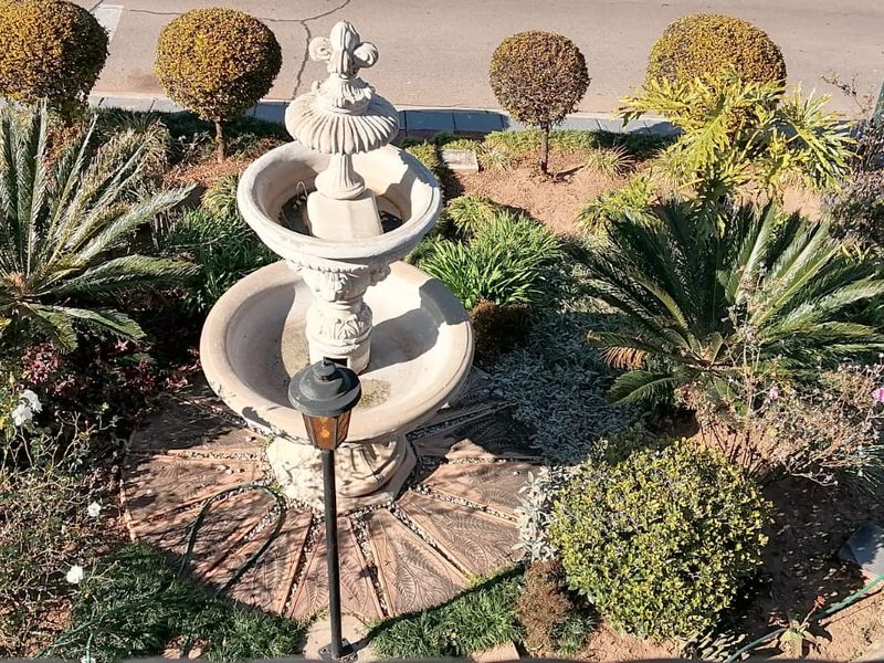 An elevated view of a landscaped garden featuring a central stone fountain, mature shrubs, and decorative brick paving.