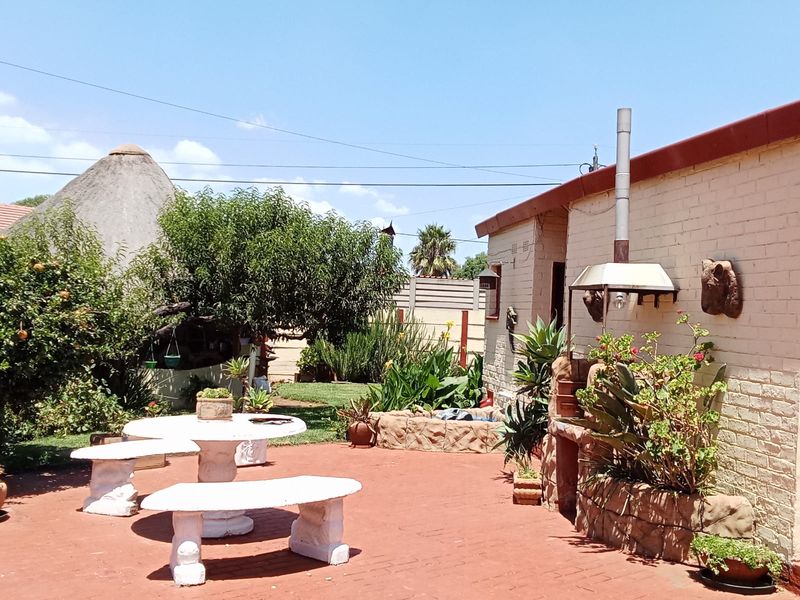 A paved outdoor patio area featuring a built-in brick braai and chimney, set within a landscaped garden with mature trees.