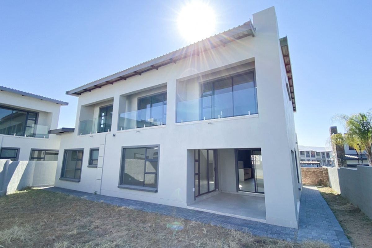 Rear view of a modern double-story house featuring a covered patio, glass balcony railings, and large aluminum-framed windows.
