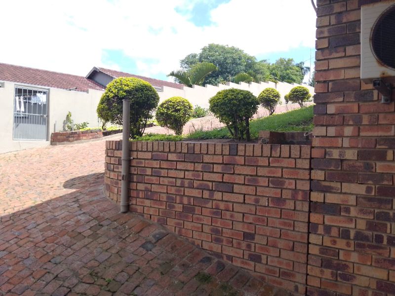 A view of the rear exterior area featuring a brick retaining wall, paved driveway, and boundary wall with established landscaping.