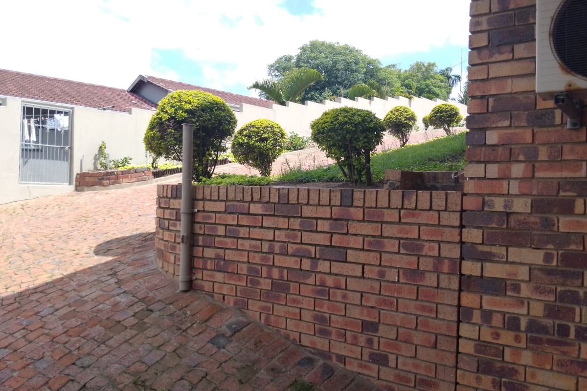 A view of the rear exterior area featuring a brick retaining wall, paved driveway, and boundary wall with established landscaping.