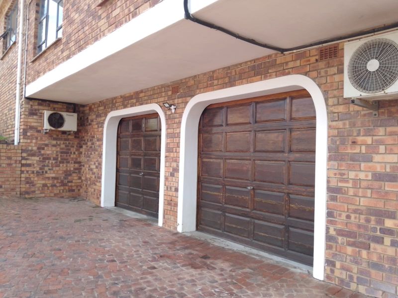 A double garage featuring traditional wooden doors, brick exterior walls, and external air conditioning units, set against a paved driveway area.