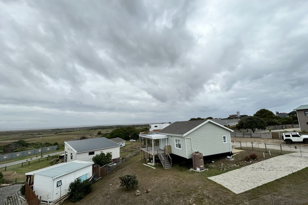 An elevated aerial view of a coastal property featuring a light-colored house with a deck, a secondary structure, and a water tank.