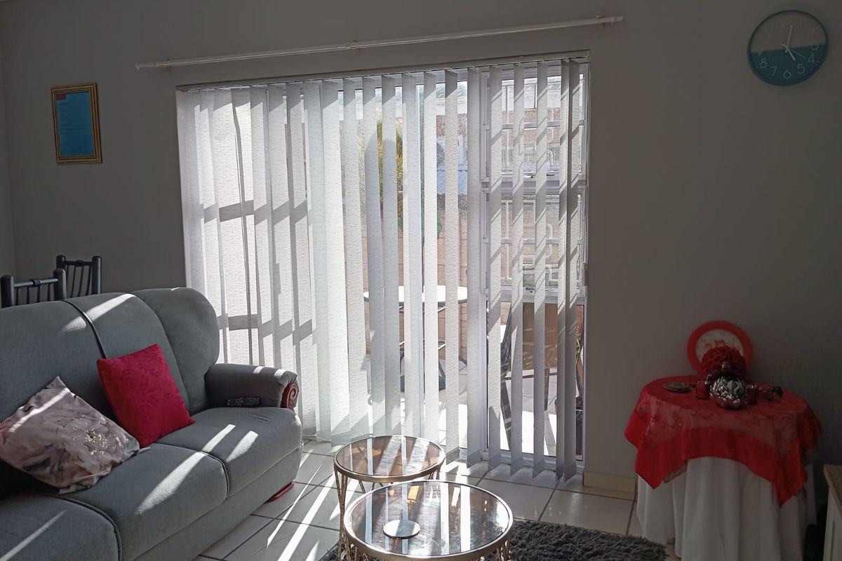 A bright living room featuring tiled flooring and large vertical blinds covering a sliding glass door that leads to an outdoor patio area.