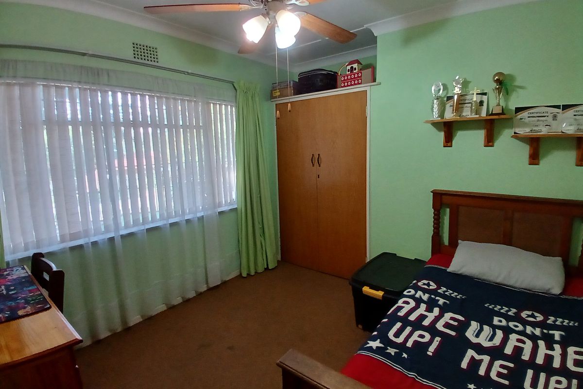 A bedroom featuring a built-in wooden wardrobe, ceiling fan, and large window with vertical blinds.