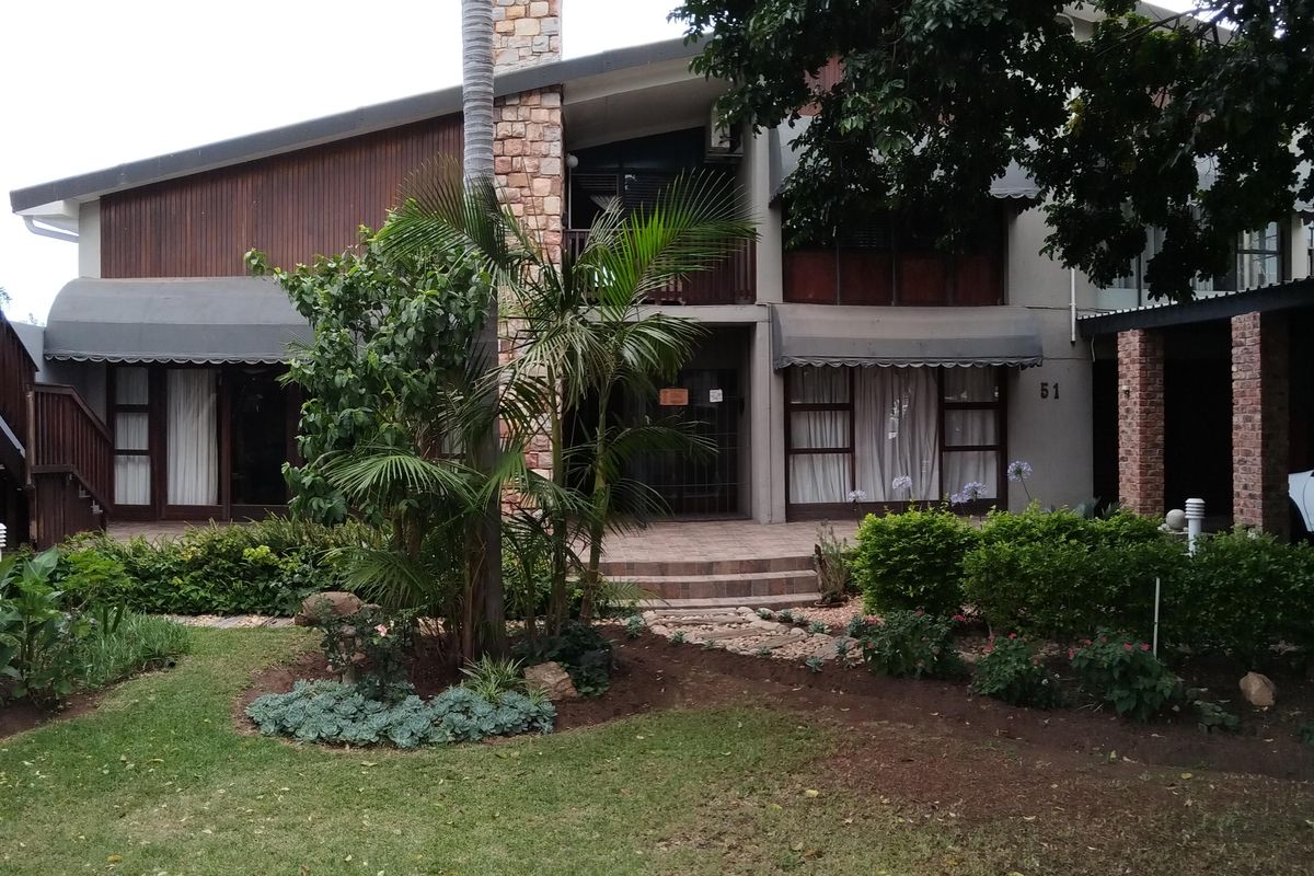 Rear view of the property featuring a stone chimney, timber cladding, and a paved patio area surrounded by established garden landscaping.