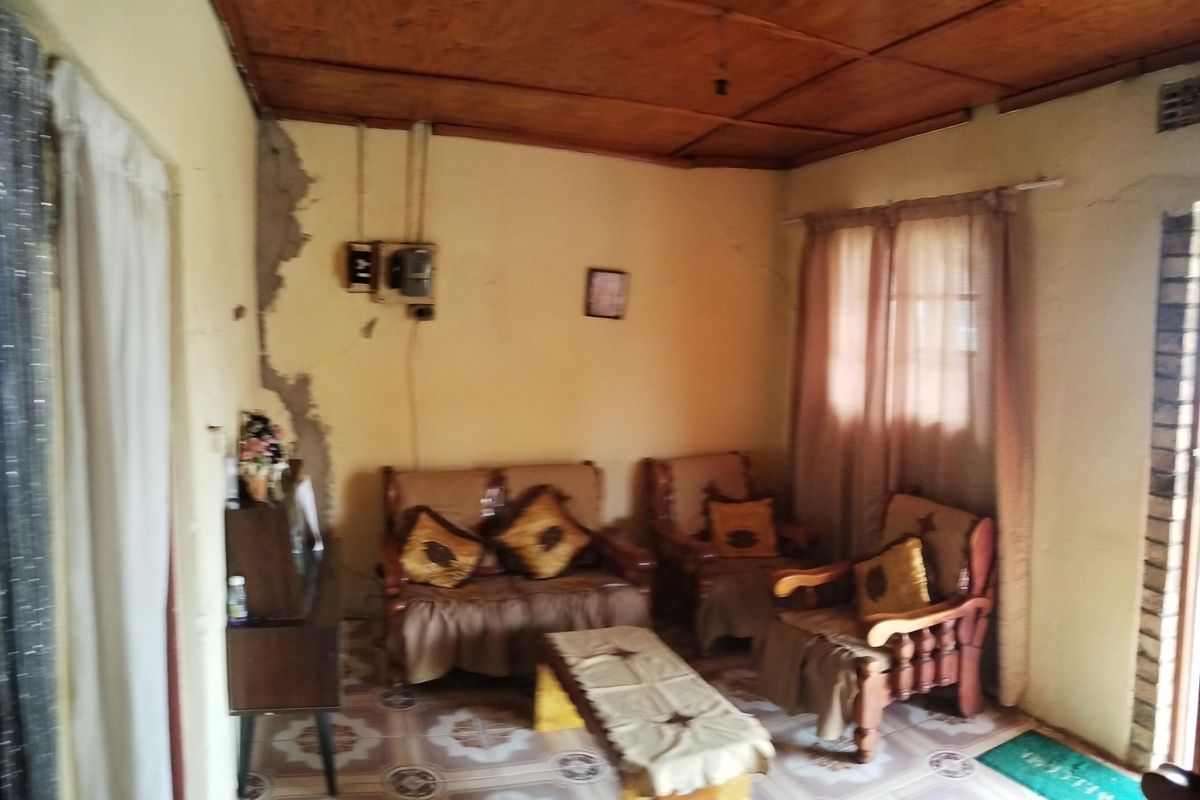 A living room featuring a wooden panel ceiling and tiled flooring, currently showing significant signs of wear and wall damage requiring maintenance.