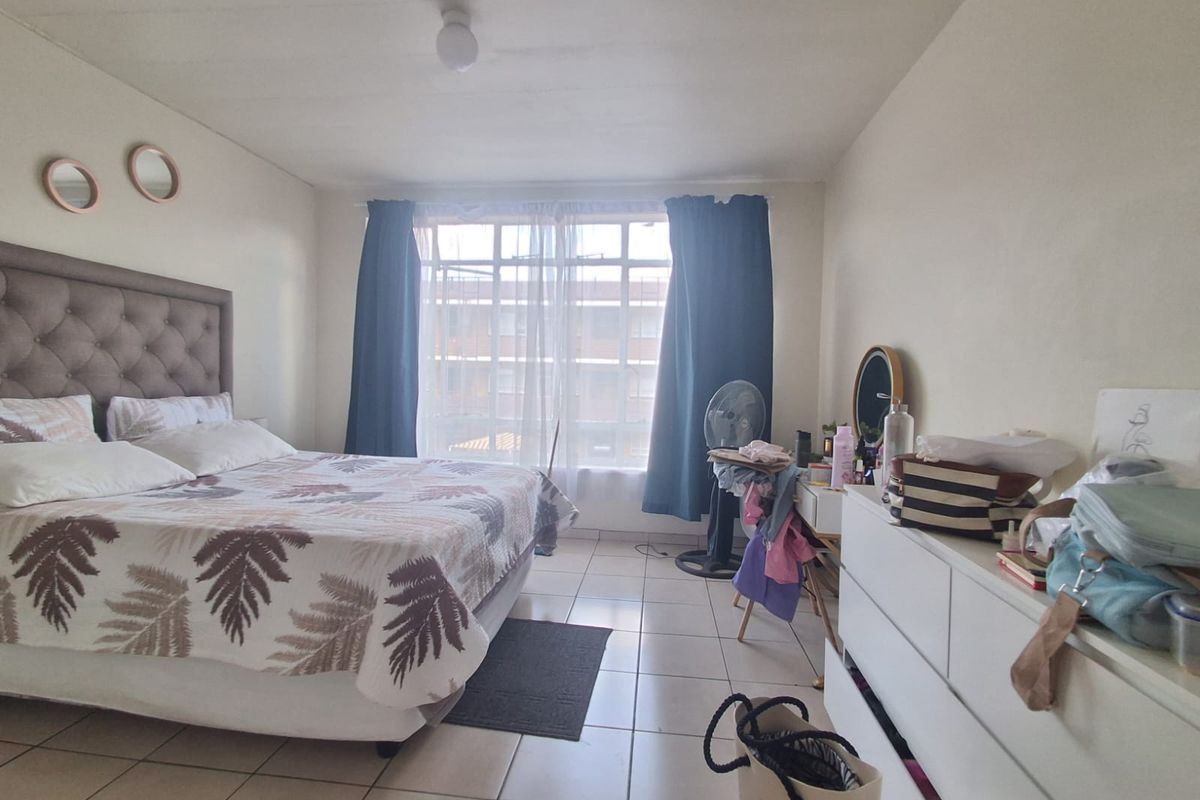 A bedroom featuring tiled flooring and a large window with curtains, providing natural light and a view of the surrounding complex.