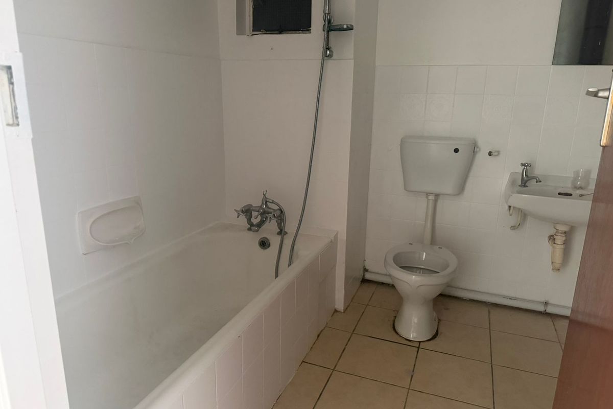 A dated bathroom featuring a standard bathtub with shower attachment, a wall-mounted toilet, and a small basin with tiled flooring and walls.