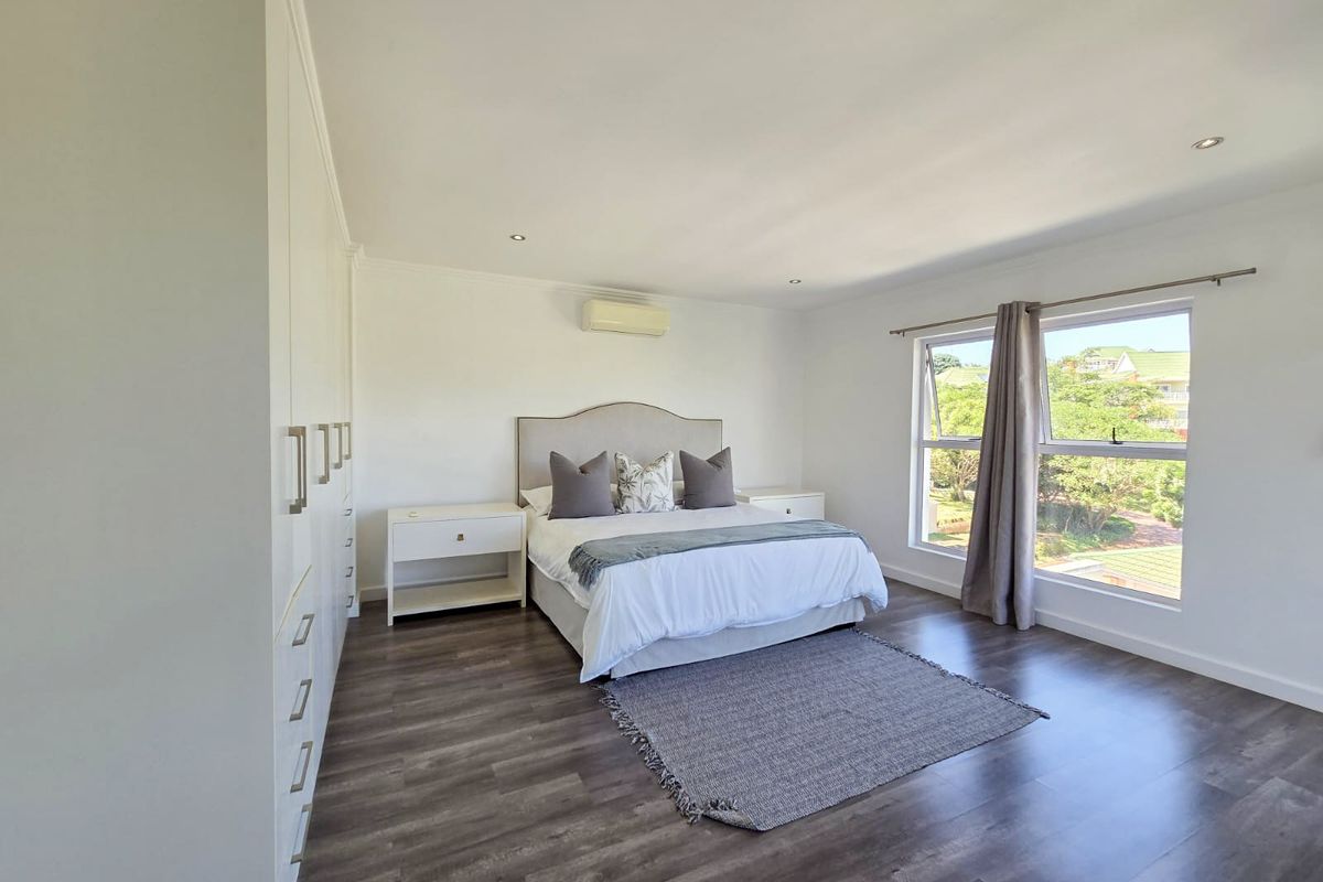 Spacious bedroom featuring built-in cupboards, laminate flooring, recessed ceiling lighting, and a wall-mounted air conditioning unit with large windows overlooking the garden.