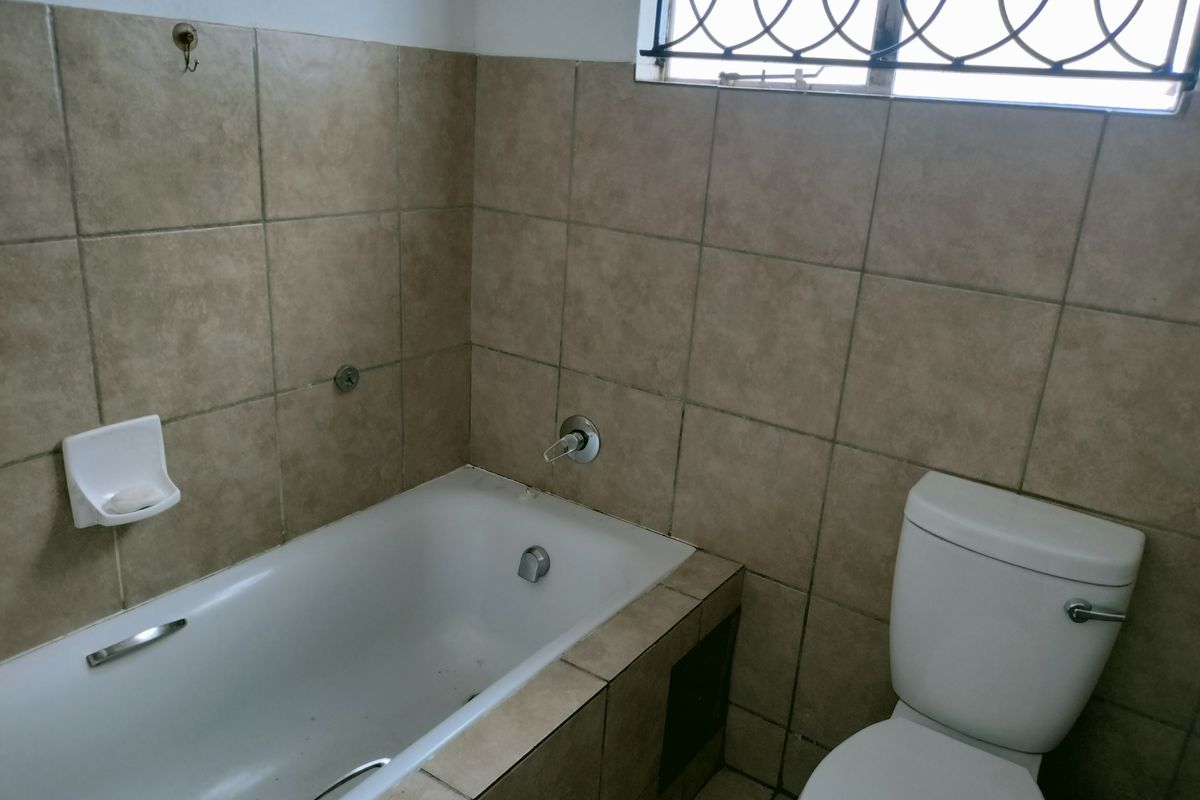 A functional bathroom featuring a built-in bathtub with tiled surround and a standard toilet, finished with neutral wall tiles and security bars on the window.