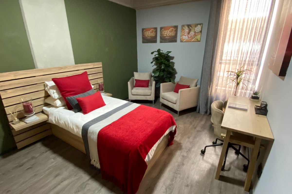 A well-lit bedroom featuring wood-look flooring, a feature wall, and large windows with sheer curtains, providing a comfortable and functional living space.