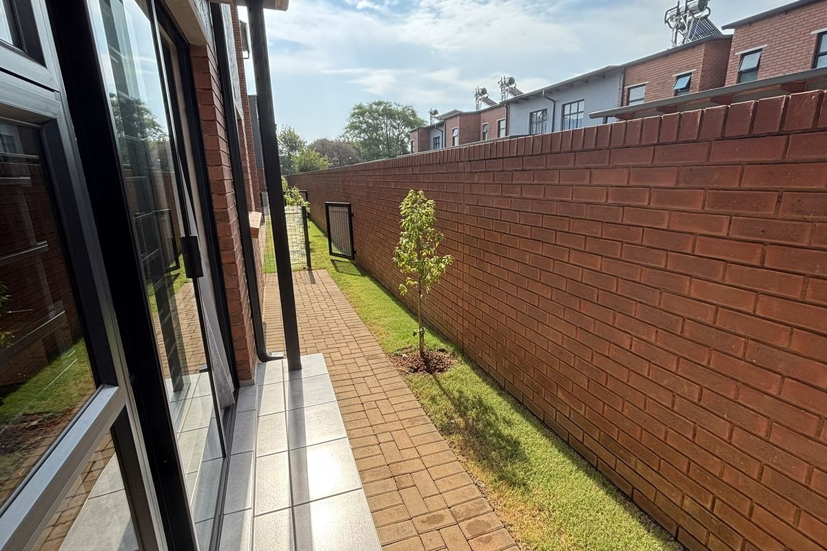 A side exterior view of the apartment featuring a paved walkway, a small lawn area, and a high brick boundary wall.