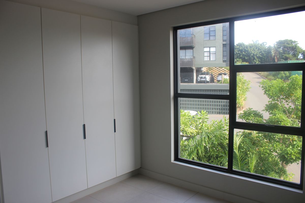 A bright bedroom featuring modern built-in cupboards and a large window with a pleasant view of the surrounding greenery and street.