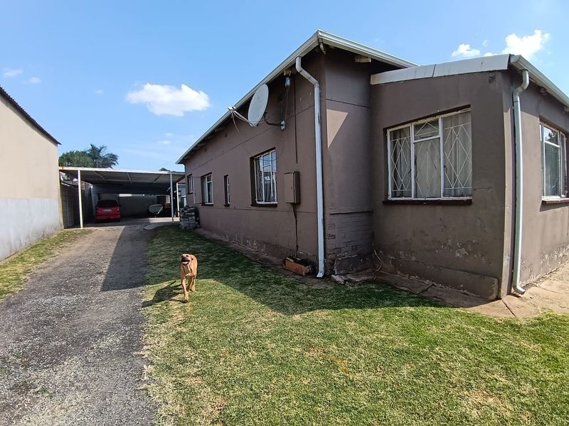 Rear exterior view of the house showing a side driveway, metal carport, and lawn area with visible wear to the exterior wall finish.