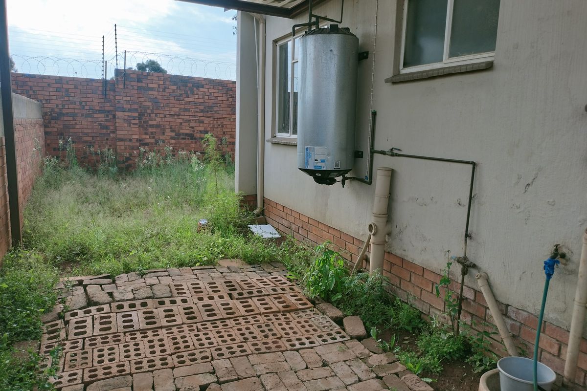 Rear exterior area featuring a wall-mounted geyser, brick paving, and an overgrown garden space enclosed by a brick boundary wall with security fencing.