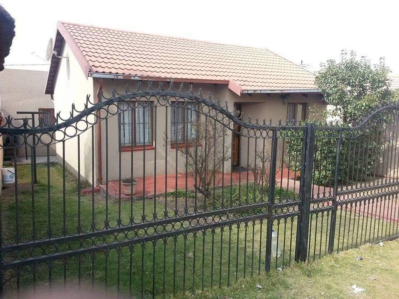 Front view of a single-story house featuring a tiled roof, paved entrance patio, and a secure wrought iron perimeter gate.