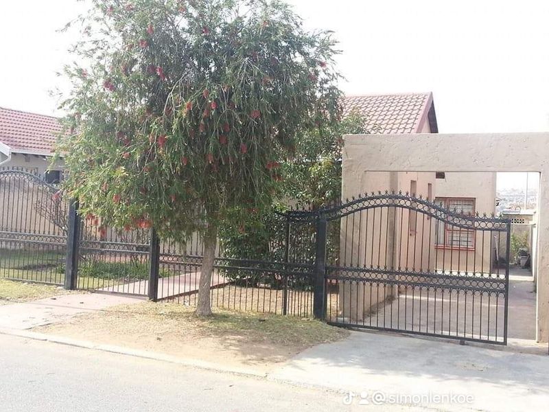 Front view of the property featuring a gated entrance, paved driveway, and established garden area with a tiled roof structure.