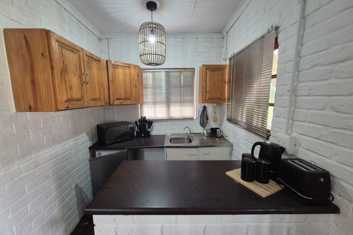 A compact kitchen featuring white painted brick walls, wooden cabinetry, a dark countertop, and a stainless steel sink.