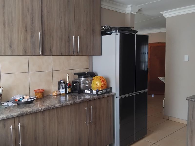A functional kitchen featuring wood-grain cabinetry, granite-look countertops, tiled backsplash, and ceramic floor tiles, leading into an adjacent hallway.