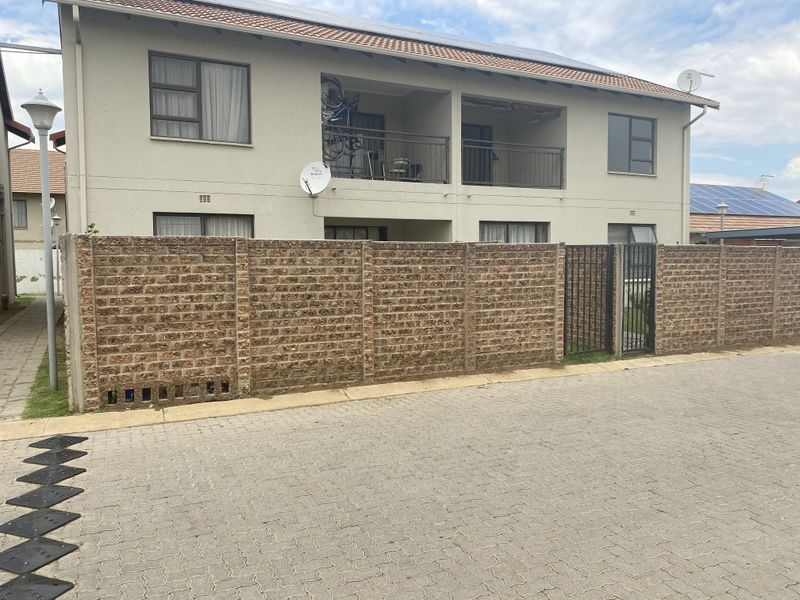 Exterior view of a townhouse featuring a brick boundary wall, paved driveway, and upper-level balcony with metal railings.