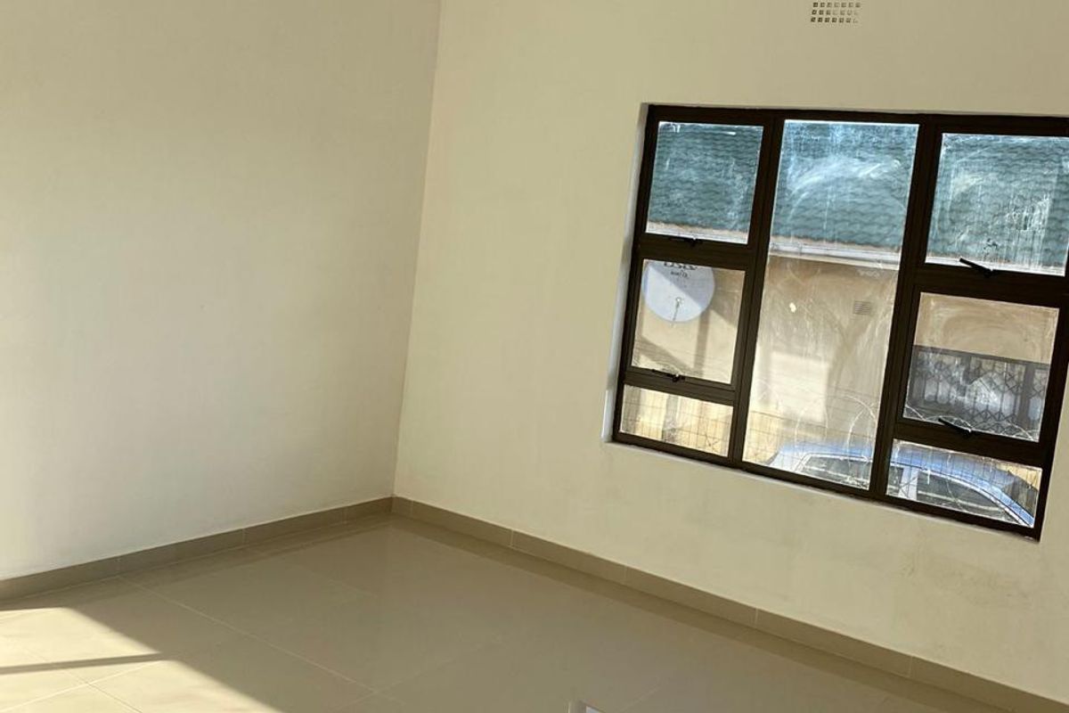 A bright, empty bedroom featuring neutral tiled flooring, white walls, and a large window with dark frames.