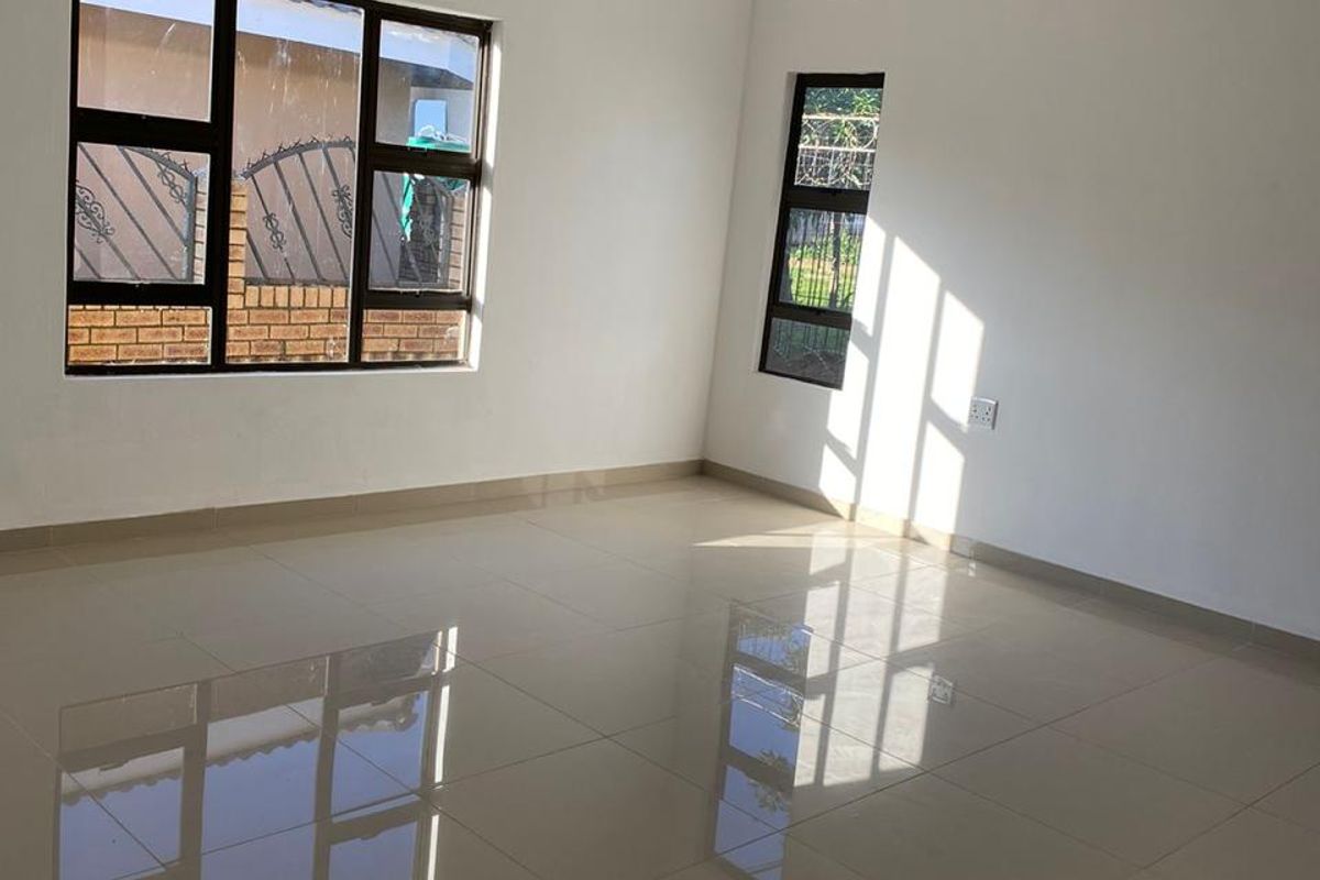 A bright, empty living room featuring polished floor tiles, white walls, decorative ceiling cornices, and large aluminium-framed windows.