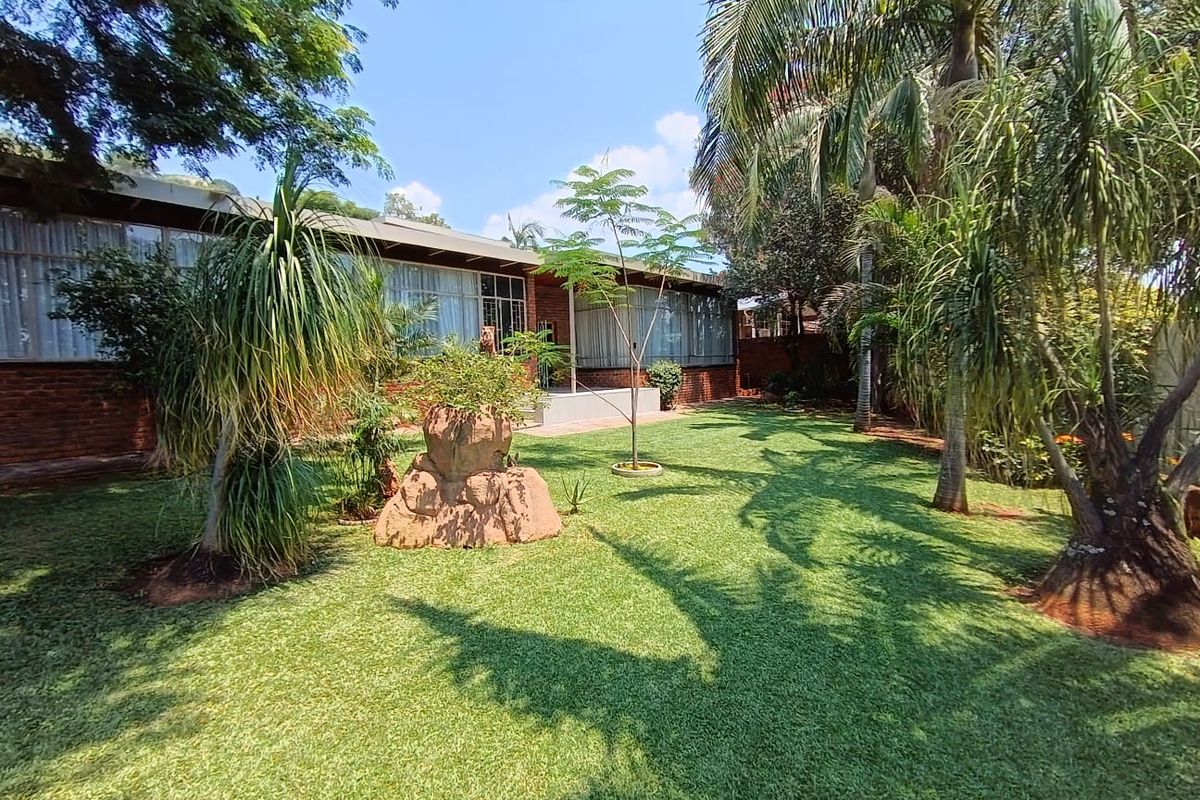 A well-maintained rear garden featuring a lush lawn, mature palm trees, and a brick-faced exterior wall with large windows.