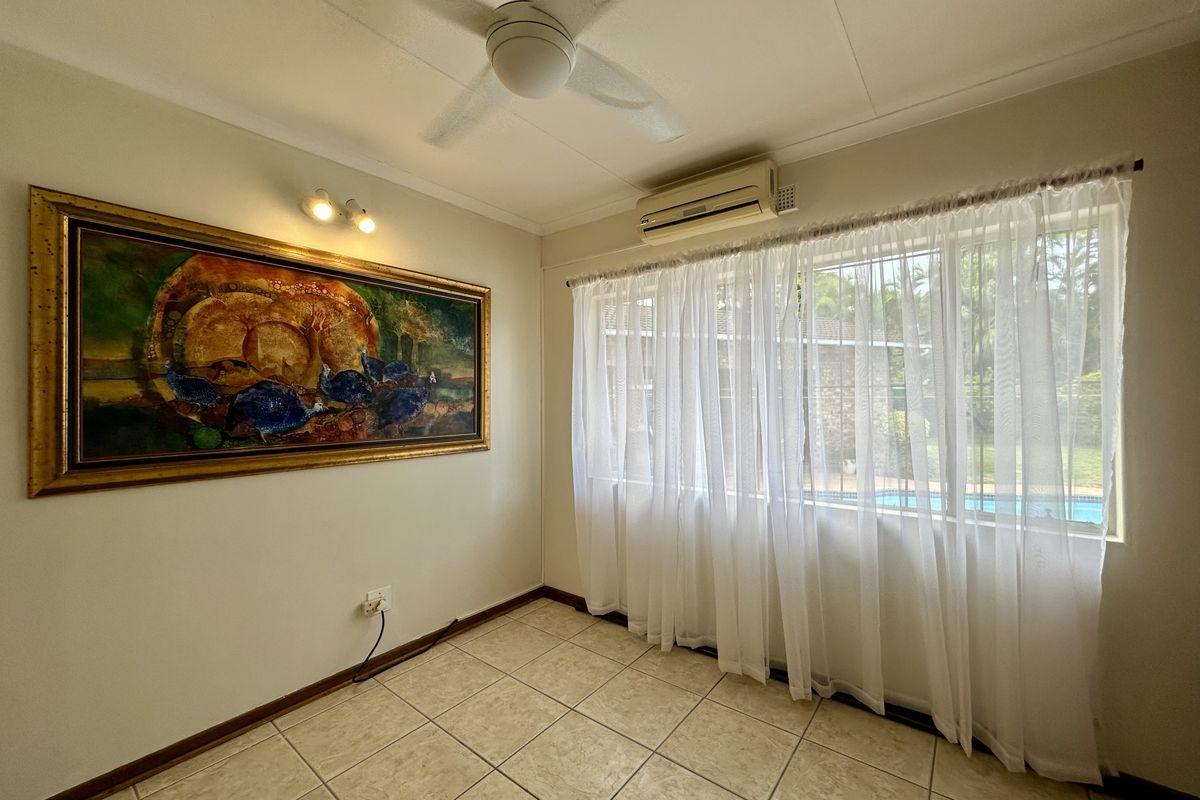 A bright bedroom featuring tiled flooring, a ceiling fan, and a wall-mounted air conditioning unit, with views overlooking the garden and pool area.