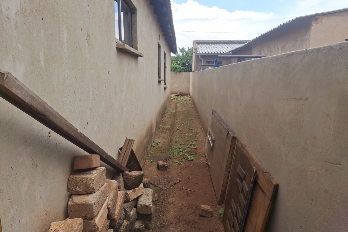 A narrow side passage between the house and boundary wall, featuring an unpaved dirt path and basic exterior wall finishes.
