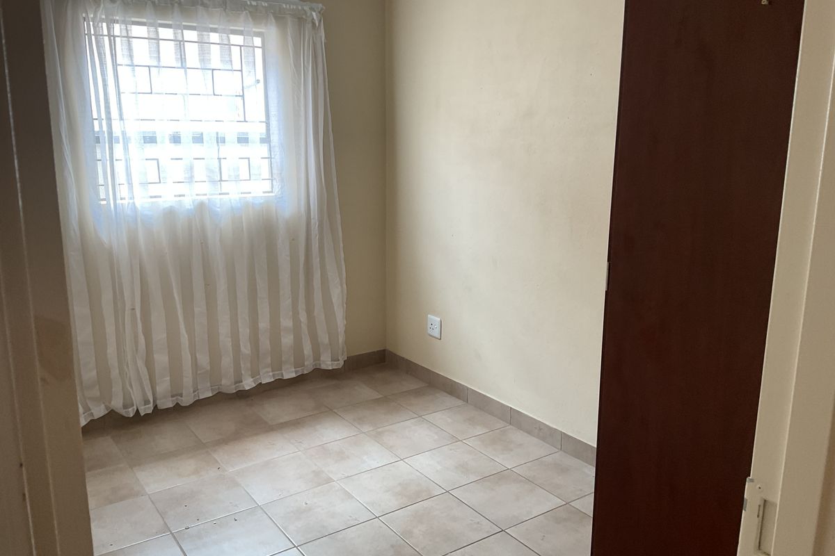 A compact, empty bedroom featuring neutral tiled flooring, a window with security bars, and a built-in cupboard.