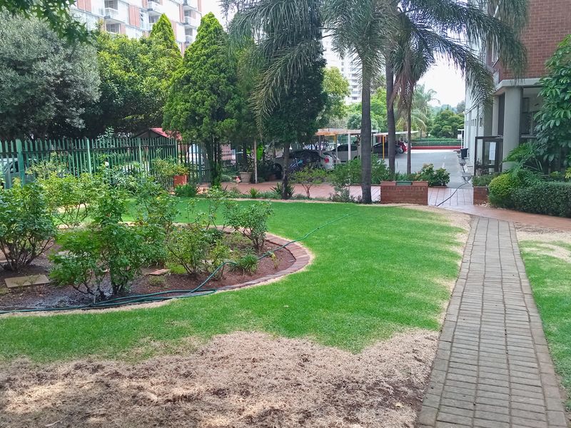 A communal garden area featuring a paved walkway, established trees, and landscaped garden beds adjacent to the apartment building.