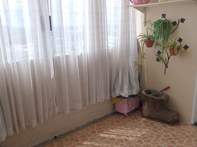 A sunlit enclosed balcony or utility space featuring tiled flooring, large windows with curtains, and wall-mounted shelving.