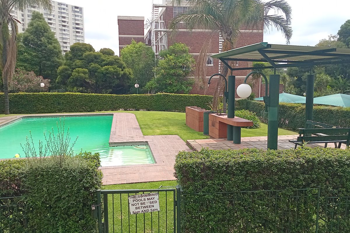 Communal outdoor pool area featuring a paved deck, manicured lawn, and a covered seating area with built-in brick braai facilities.