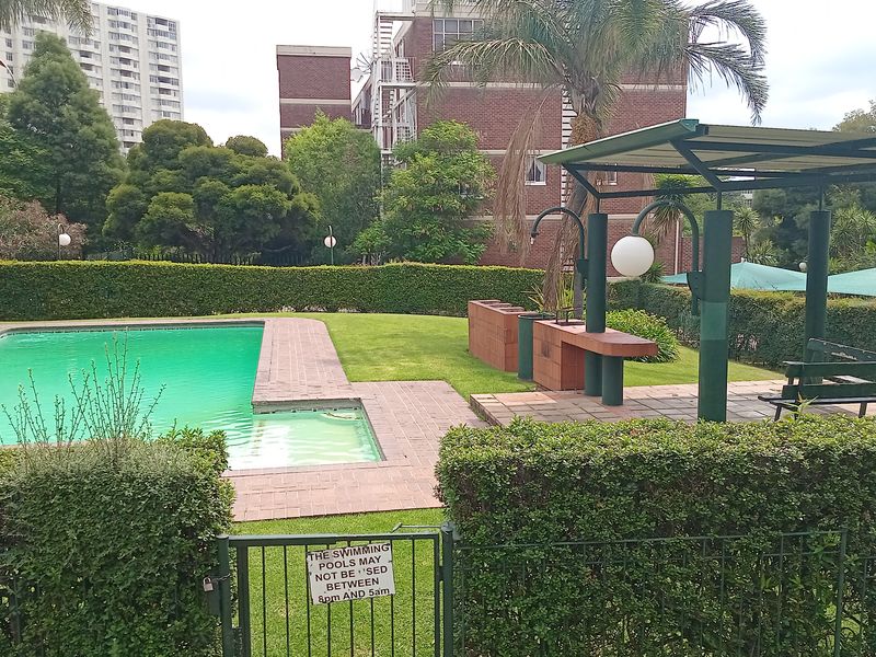 Communal outdoor pool area featuring a paved deck, manicured lawn, and a covered seating area with built-in brick braai facilities.