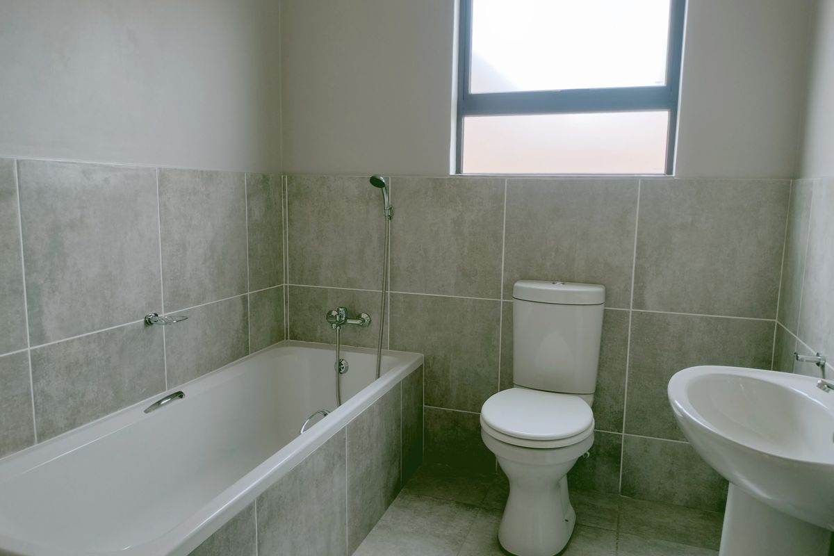 A clean, modern bathroom featuring grey wall and floor tiles, a white bathtub with handheld shower, a pedestal basin, and a toilet.