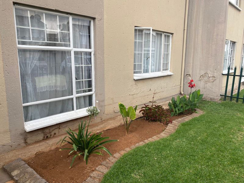 Front exterior view of the townhouse featuring a small garden bed, lawn, and security-barred windows with visible plaster damage on the exterior wall.