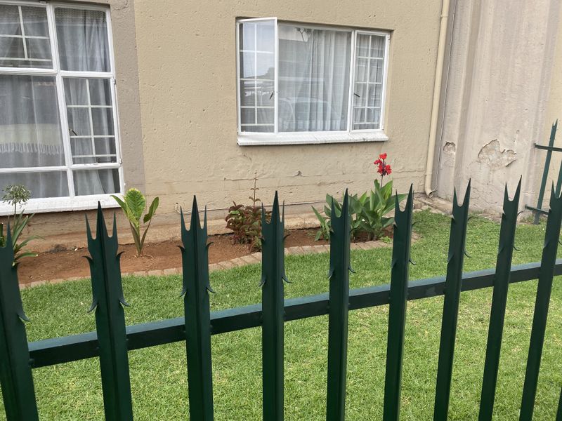 Exterior view of the townhouse facade featuring metal security fencing, a small garden bed, and aluminium window frames with visible wall plaster damage.